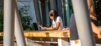 A student sitting outdoors at a desk simultaneously working on their laptop and mobile phone 